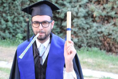 Mezuniyet kıyafetleri içinde, sarılmış diploması ve parkta piposu olan yakışıklı bir gencin yakın plan portresi.