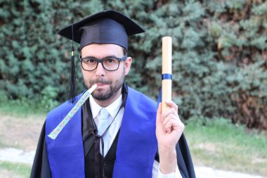 Mezuniyet kıyafetleri içinde, sarılmış diploması ve parkta piposu olan yakışıklı bir gencin yakın plan portresi.