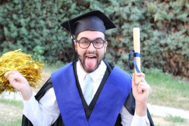 Mezuniyet kıyafetleri içinde yakışıklı genç bir öğrencinin yakın plan portresi. Diplomalı ve parkta altın bir ponpon.