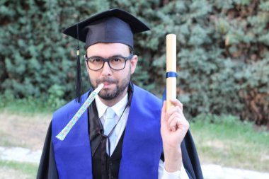 Mezuniyet kıyafetleri içinde, sarılmış diploması ve parkta piposu olan yakışıklı bir gencin yakın plan portresi.