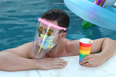 Coronavirus tecridi sırasında yüzme havuzunda gökkuşağı renkli kağıt bardakla tıbbi koruyucu maskeli yakışıklı genç adamın yakın plan portresi.