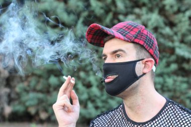 Sağlık maskesi takan yakışıklı bir gencin portresi. Ağzı delikli, yeşil parkta sigara içen, komedi anlayışı.