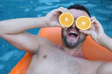 Yüzme havuzunda portakal yarımları tutan, şişme yatakta yüzen yakışıklı genç adamın yakın plan portresi