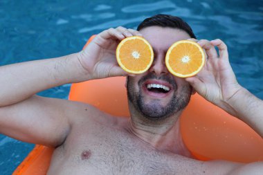 Yüzme havuzunda portakal yarımları tutan, şişme yatakta yüzen yakışıklı genç adamın yakın plan portresi