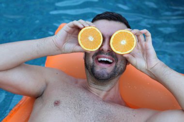 Yüzme havuzunda portakal yarımları tutan, şişme yatakta yüzen yakışıklı genç adamın yakın plan portresi