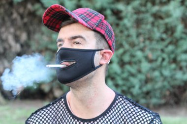 Sağlık maskesi takan yakışıklı bir gencin portresi. Ağzı delikli, yeşil parkta sigara içen, komedi anlayışı.