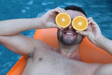 Yüzme havuzunda portakal yarımları tutan, şişme yatakta yüzen yakışıklı genç adamın yakın plan portresi