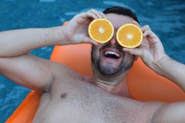 Yüzme havuzunda portakal yarımları tutan, şişme yatakta yüzen yakışıklı genç adamın yakın plan portresi