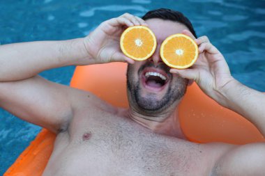Yüzme havuzunda portakal yarımları tutan, şişme yatakta yüzen yakışıklı genç adamın yakın plan portresi
