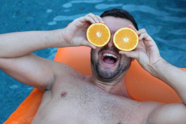 Yüzme havuzunda portakal yarımları tutan, şişme yatakta yüzen yakışıklı genç adamın yakın plan portresi