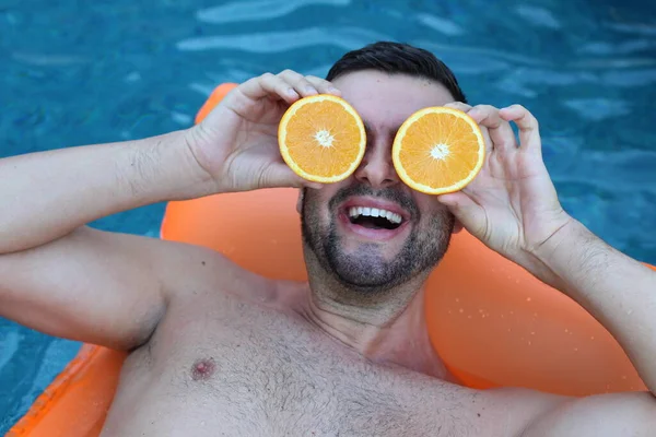 Yüzme havuzunda portakal yarımları tutan, şişme yatakta yüzen yakışıklı genç adamın yakın plan portresi