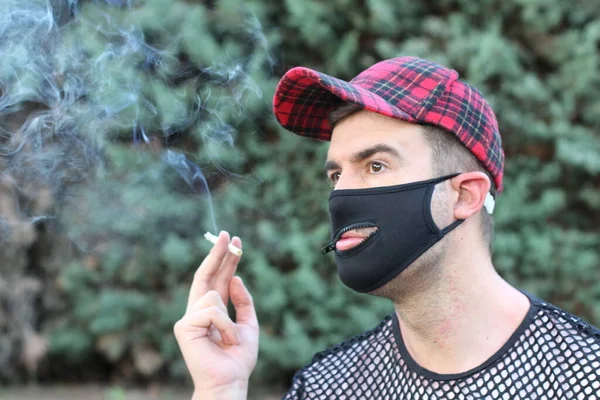 閉鎖ポートレートのハンサムな若いです男とともに医療マスクのためのCovid保護のための穴のための口の喫煙で緑の公園 コメディコンセプト — ストック写真