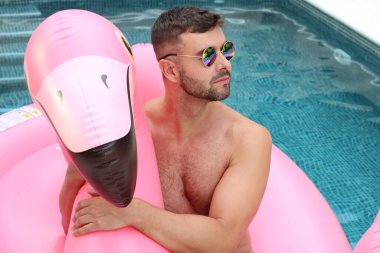 Yüzme havuzunda pembe şişme flamingo ile kameraya bakan yakışıklı genç adamın yakın plan portresi.