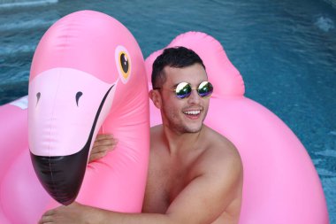 Yüzme havuzunda pembe şişme flamingo ile kameraya bakan yakışıklı genç adamın yakın plan portresi.
