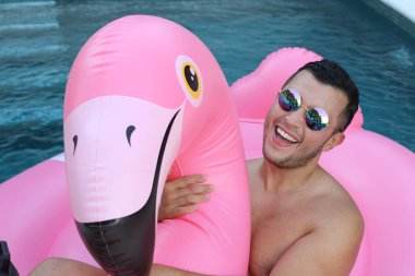 Yüzme havuzunda pembe şişme flamingo ile kameraya bakan yakışıklı genç adamın yakın plan portresi.
