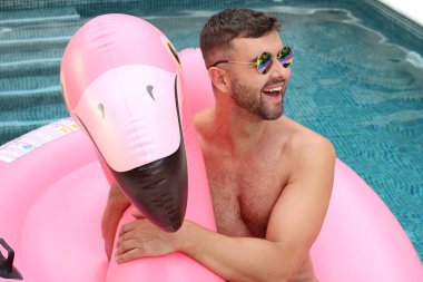 Yüzme havuzunda pembe şişme flamingo ile kameraya bakan yakışıklı genç adamın yakın plan portresi.
