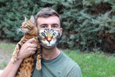 Nefes maskeli yakışıklı genç adamın yakın plan portresi. Kedi suratlı, tekir kedisini açık havada tutuyor ve kameraya bakıyor.