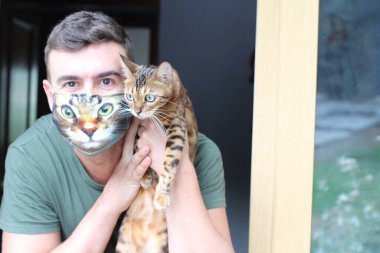 Nefes maskeli yakışıklı genç adamın yakın plan portresi. Kedi suratlı, tekir kedisini açık havada tutuyor ve kameraya bakıyor.