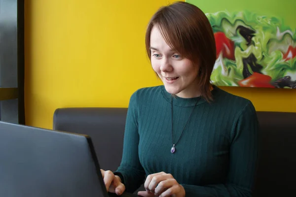 Menina com uma expressão tensa surpreso perplexo senta-se trabalhando em um laptop em uma mesa em um café em um sofá marrom contra uma parede amarela — Fotografia de Stock