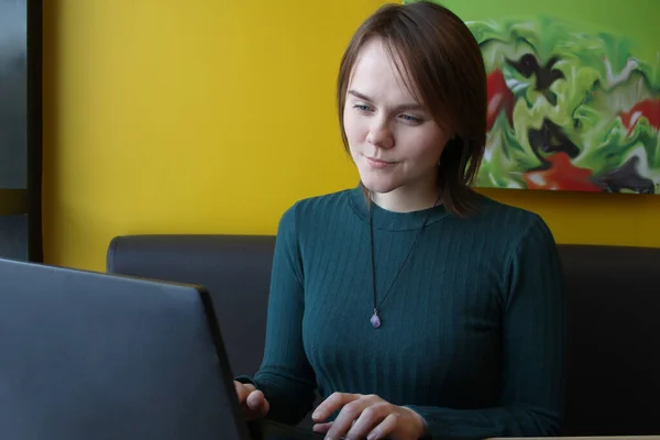 Uma menina com uma expressão tensa pensiva interessada em seu rosto se senta, trabalhando em um laptop em uma mesa em um café em um sofá marrom contra uma parede amarela. Ele olha para a tela do computador com olhos estreitos — Fotografia de Stock