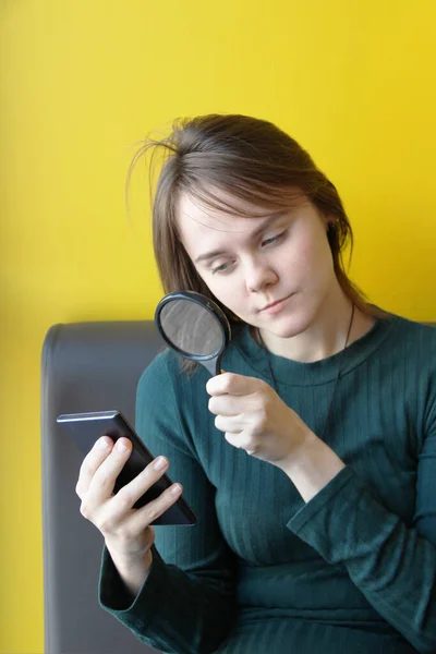 Una joven hermosa chica de aspecto europeo en una chaqueta azul se sienta en un café con un teléfono móvil en el sofá contra el fondo de una pared amarilla. Mira a través de una lupa en los aparatos —  Fotos de Stock