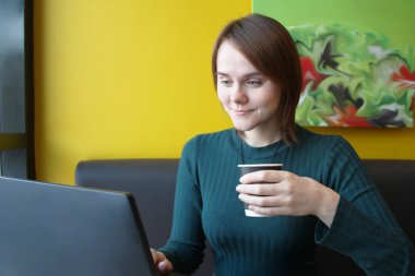 Yüzünde neşeli bir ifade olan bir kız sarı bir duvara karşı kahverengi bir kanepede dizüstü bilgisayarda otururken gülümsüyor. Elinde kahve olan bir kağıt bardağı tutan bir bilgisayar monitörüne bakıyor..