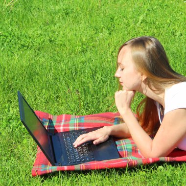 Beyaz tişörtlü, uzun saçlı, güzel beyaz bir kız kırmızı ekoseli, yeşil çimenli, çimenli ve siyah bir dizüstü bilgisayarın arkasında çalışıyor..