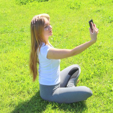 Beyaz tişörtlü güzel beyaz bir kız yeşil çimlerin üzerinde oturan uzun saçlı ve selfie çeken..
