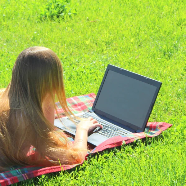 Beyaz tişörtlü, uzun saçlı, güzel beyaz bir kız kırmızı ekoseli, yeşil çimenli, çimenli ve siyah bir dizüstü bilgisayarın arkasında çalışıyor..