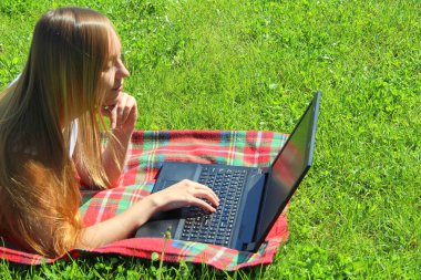 Beyaz tişörtlü, uzun saçlı, güzel beyaz bir kız kırmızı ekoseli, yeşil çimenli, çimenli ve siyah bir dizüstü bilgisayarın arkasında çalışıyor..