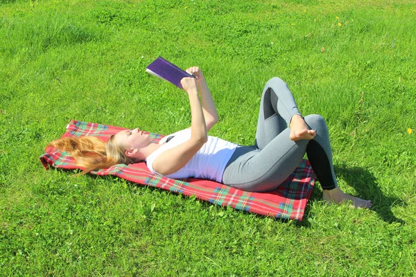 Piękna biała dziewczyna w białym T-shircie z długimi włosami leży na czerwonej kratę, na zielonej trawie, na trawniku i czytać książki — Zdjęcie stockowe