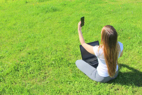 Una hermosa joven blanca en una camiseta blanca y con el pelo largo sentado en la hierba verde, en el césped y trabajando detrás de un ordenador portátil negro con un teléfono móvil y toma una selfie — Foto de Stock