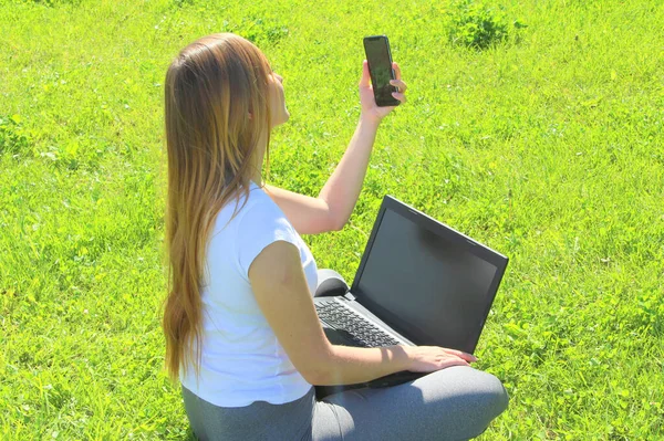 Una hermosa joven blanca en una camiseta blanca y con el pelo largo sentado en la hierba verde, en el césped y trabajando detrás de un ordenador portátil negro con un teléfono móvil y toma una selfie — Foto de Stock