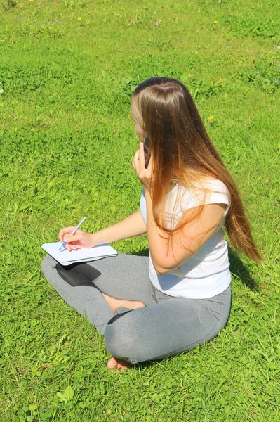 Una hermosa joven blanca en una camiseta blanca y con el pelo largo sentado en la hierba verde, en el césped, hablando en un teléfono celular y escribe con una pluma en un cuaderno —  Fotos de Stock