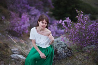 Bir kız çiçek açmış bir şekilde bir dağ manzarasının arka planında yürür. Altai dağlarında ledum çiçekleri açar..