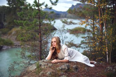 Peri masalı. Nehir perisinin hikayesi. Altai 'de ateş etmek. Beyaz elbiseli bir kız nehir kıyısındaki kayalık bir sahilde yürüyor..