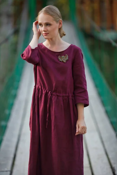 Girl Burgundy Dress Walks Suspended Wooden Bridge Smiling Blonde — Stock Photo, Image