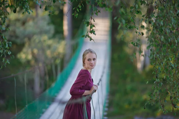 Girl Burgundy Dress Walks Suspended Wooden Bridge Smiling Blonde — Stock Photo, Image