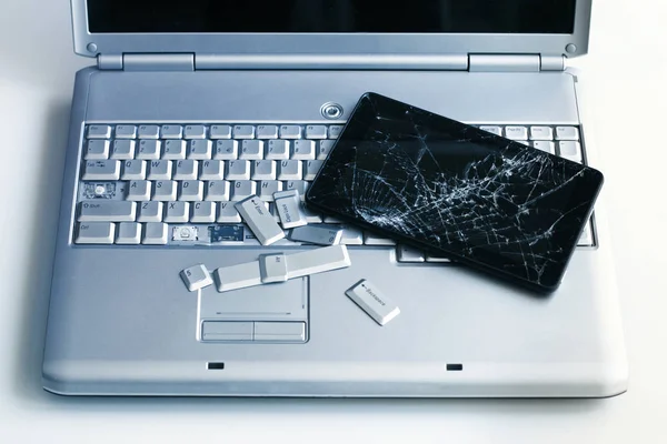 A silver laptop with a broken keyboard and a tablet with a cracked display. A close-up picture of part of broken laptop and cracked screen on a tablet.