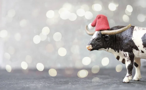 toy ox or bull in red santa hat as symbol of 2021 year. chinese calendar or horoscope. new year card or poster. bull on grey defocused background with bokeh. lights. happy new year concept.