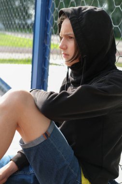 Spor parkında çitlerin yanında oturan yakışıklı genç çocuk. Başlığı kafasında siyah kapüşonlu bir genç. Üzgün adam yalnız oturuyor.. 
