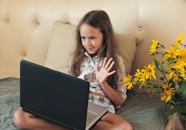 A girl communicate with friend on-line by video call on laptop. schoolgirl using videoconferencing app. video chat concept. a girl sitting on a bed and waving hand to a teacher. online education.