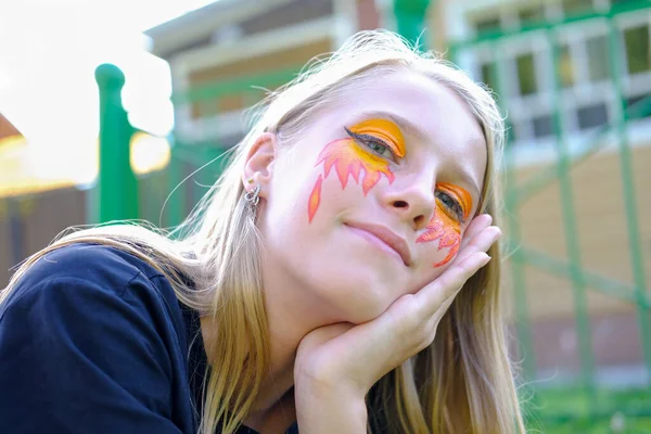 Beautiful teenager girl with face painting lying on a lawn. smiling blonde girl with fire and flame festive face painting. fire face art on teenager girl\'s eyes and cheeks.