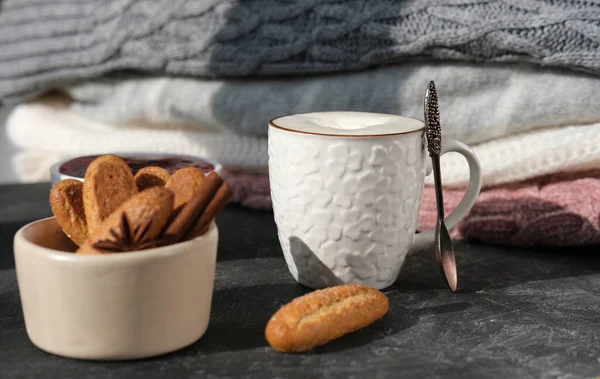 white cup of coffee and sugar cookies near a pile of knitted sweaters. autumn sunny morning. fall season mood. warm sweaters and coffee cup on a grey table.