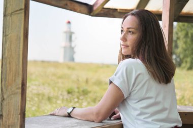 Uzun kahverengi saçlı güzel bir kadın. Bir tarafa bakıyor ve düşünüyor. Kırsal kesimde ahşap bir çadırda duran düşünceli orta yaşlı kadın. Arka planda bir deniz feneri.