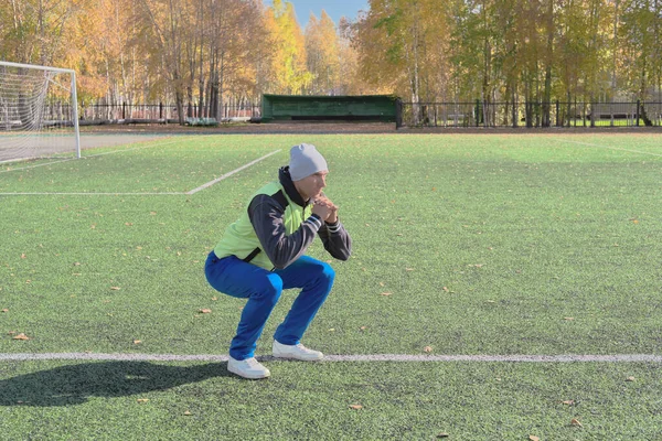 handsome caucasian man doing squats on a stadium football field. fall season open air workout. toned sporty man doing exercises outdoors.