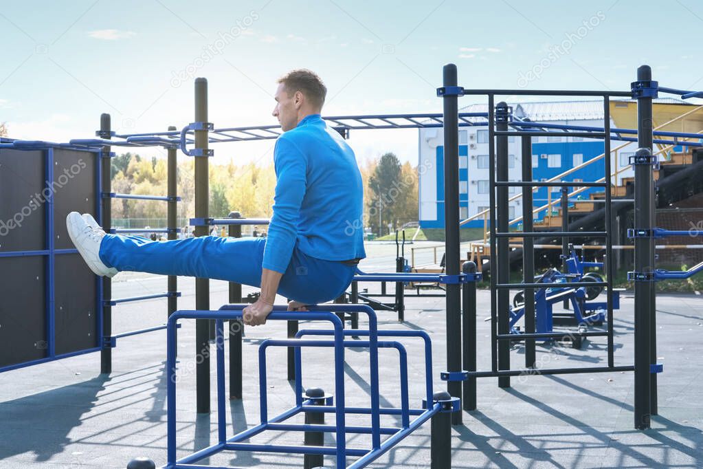 sportsman of middle age working out on a stadium open air gym. L-sit or legs raise on a parallel bars. active lifestyle. sunny autumn day on a stadium. open air training equipment.