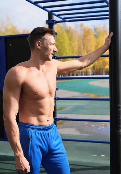 middle age shirtless man resting after outdoors workout. autumn season training. outdoors stadium. ladders and monkey bars for phisical activity. toned man with strong abs looking aside