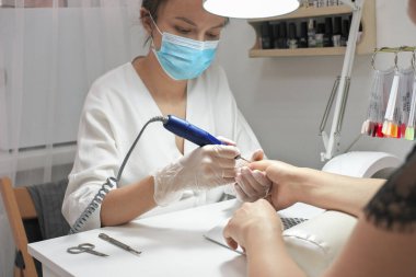 Genç bayan manikürcü müşteri için manikür yapıyor. Güzellik uzmanı ojeye oje hazırlamak için oje makinesi kullanıyor. Koruyucu maskeli kadın güzellik salonunda çalışıyor..