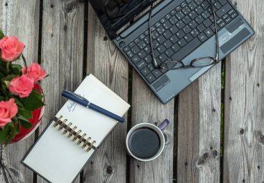 Bahçedeki ahşap bir masada üzerinde kahve ve not defteri olan dizüstü bilgisayar, evden çalıştığını gösteriyor.